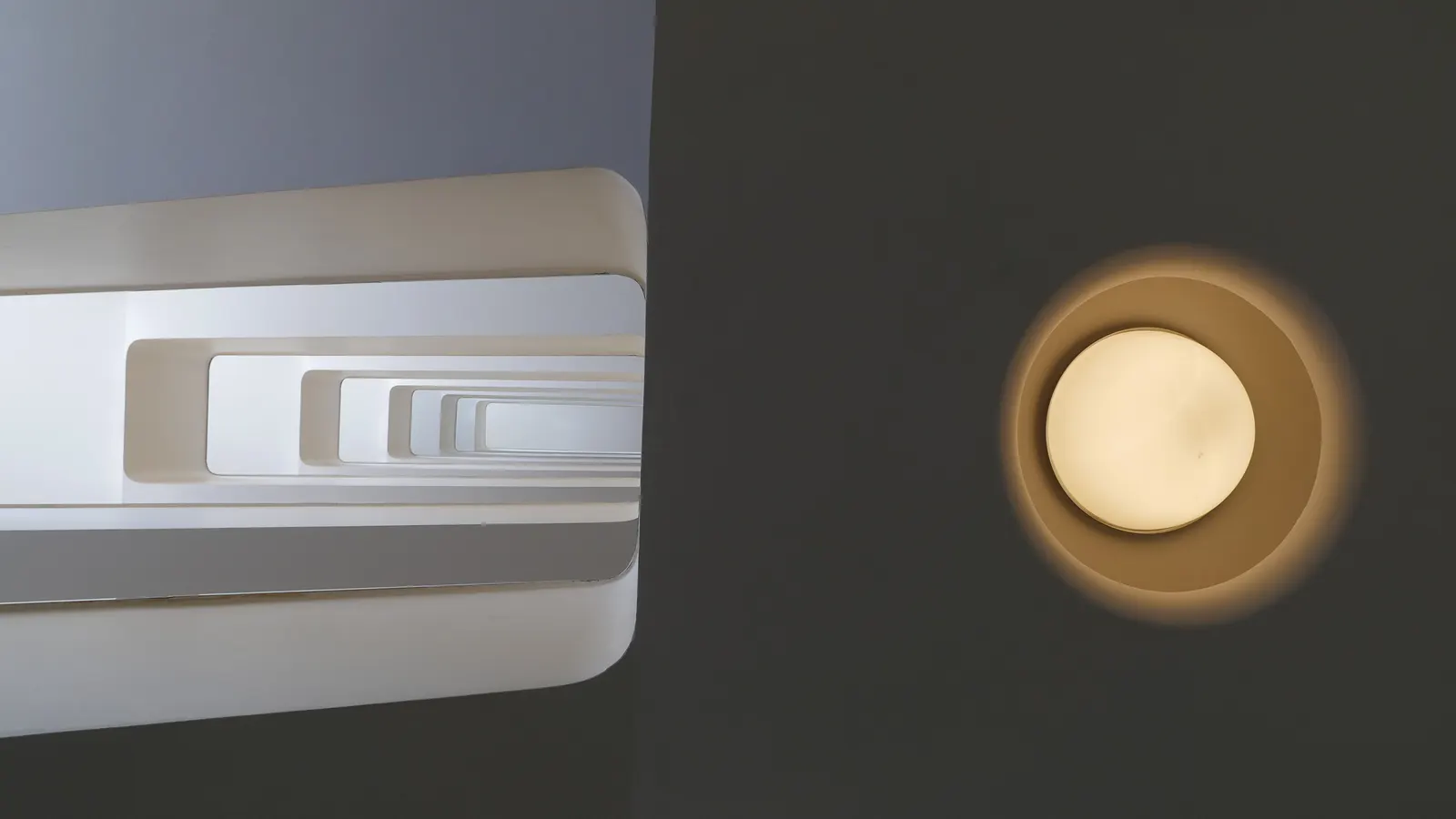 Cage d'escalier photographiée d'en bas, à côté d'un plafonnier rond sur un mur sombre